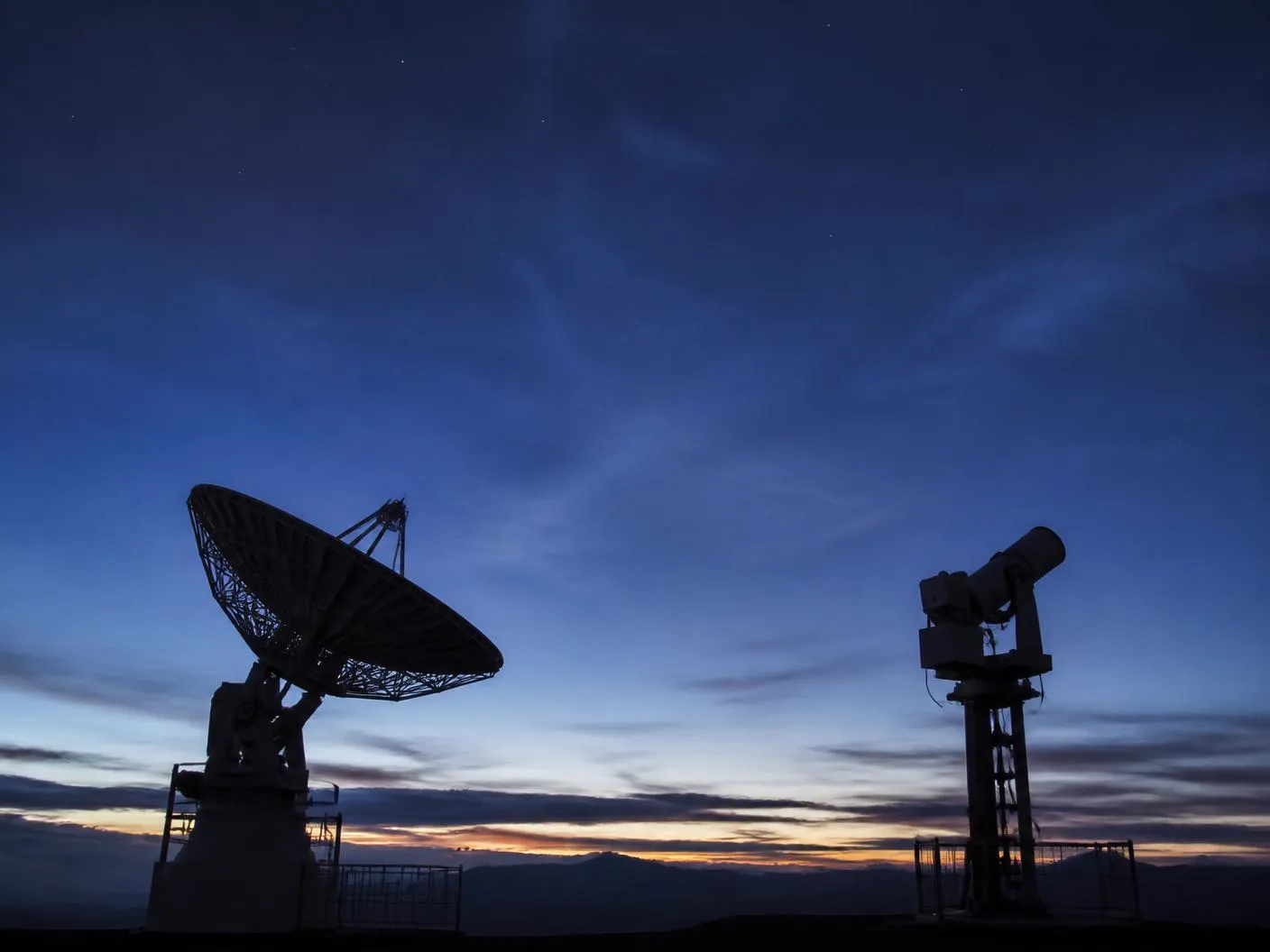 Ground-based radar dish and optical tracking instrument silhouetted against a twilight sky
